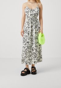Robe maxi à imprimé floral en crème et noir, avec des bretelles fines, une taille nouée, associée à des sandales à plateforme épaisses noires et un sac vert duveteux.