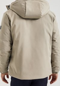 Béžová bunda s kapucí a elastickými manžetami, kterou má na sobě osoba otočená zády, v kombinaci s tmavými kalhotami na jednobarevném pozadí.