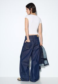 Teksas hlače z visokim pasom, širokega kroja v temno modri barvi, z velikimi žepi in kontrastnim šivom, kombinirane s prilegajočo belo kratko majico.