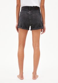 Person som står barfota, iförd uppvikta svarta jeansshorts och en vit topp, sedd bakifrån mot en enkel vit bakgrund.