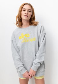 Grauer Sweatshirt mit langen Ärmeln und gerippten Bündchen. Mit gelber Grafikschrift, die "elko Freestyle" sagt. Weicher, lässiger Schnitt.