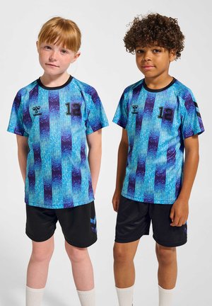 Zwei Kinder tragen passende blau-lila gestreifte Sporttrikots mit der Nummer 12 und schwarze Shorts vor einem einfarbigen Hintergrund.