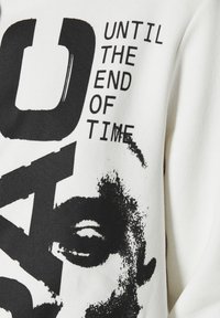 Sudadera blanca con texto gráfico negro en negrita e ilustración de una cara. El texto dice "HASTA EL FINAL DE LOS TIEMPOS". Material de algodón suave.