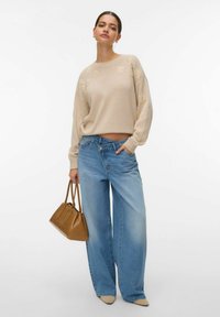 Femme portant un pull beige en maille à motifs floraux et un jean à jambes larges asymétriquement boutonné, tenant un sac à main marron, debout devant un fond blanc.
