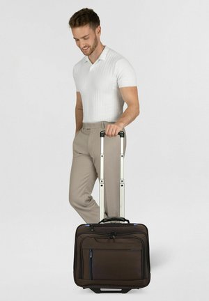 Uomo con polo bianca e pantaloni beige che tiene una borsa per laptop con ruote per manico esteso, guarda verso il basso e sorride.