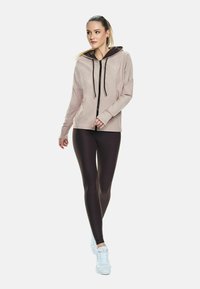 Young woman walking, wearing light beige zip-up hoodie, black leggings, and white sneakers, with hair tied back and neutral expression.