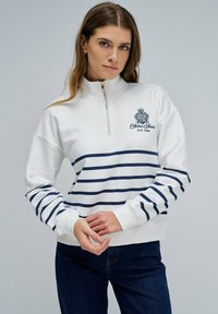 Sudadera blanca con cremallera y rayas horizontales en azul marino, con un logo bordado en el pecho. Cuenta con puños acanalados y un diseño de cuello alto.