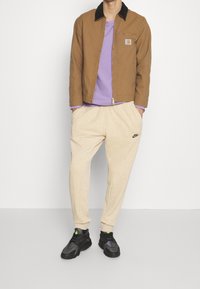 Ruskea takki mustalla kauluksella, violetti paita, kermaiset collegehousut ja mustat lenkkarit vihreillä yksityiskohdilla. Yksinkertainen muotoilu, rento istuvuus.