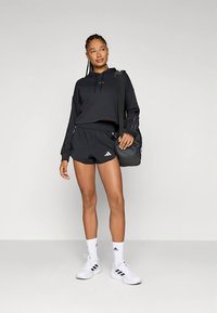 Jovem mulher com um moletom atlético preto e calções, usando sapatilhas e meias brancas da Adidas, segura uma mala preta de ombro, enquanto está em pé contra um fundo simples.