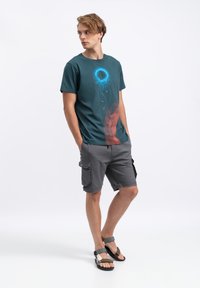 Teal katoenen t-shirt met een cirkelvormige blauwe afbeelding, rode accenten; gecombineerd met grijze cargo shorts en zwarte sandalen met verstelbare banden.