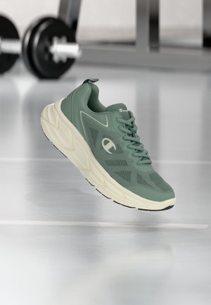 Grüner und beiger Sportschuh mit Mesh-Obermaterial und klobiger Sohle, der über einem reflektierenden Fitnessboden mit Gewichthebergeräten im Hintergrund schwebt.