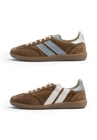 Deux baskets en daim marron avec semelles en gomme, l'une avec des rayures bleues et un patin de talon, l'autre avec des rayures blanches et un patin de talon, vues de côté.
