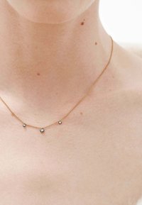 Collar de oro con tres pequeñas gemas transparentes y redondas. Cadena delicada con acentos espaciados uniformemente, llevada en un cuello descubierto.