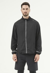 Svart lättviktsjacka med ståkrage, vita accentlinjer, dragkedja och sidfickor, kombinerad med svarta shorts.