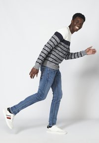 Gestreepte trui in marineblauw, grijs en wit, gecombineerd met lichtblauwe spijkerbroek en witte sneakers met zwarte accenten. Model wandelt casual.