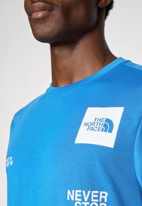 Blå bomull t-skjorte med korte ermer. Har et hvitt logo-patch og tekst på forsiden; rund hals design. Glatt tekstur.