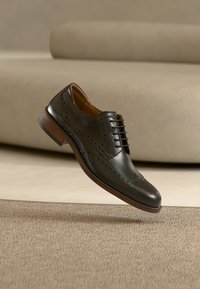 Μαύρο δερμάτινο παπούτσι brogue με ξύλινη σόλα, λεπτομερείς διάτρητες λεπτομέρειες και κορδόνια, αιωρούμενο πάνω από μπεζ χαλί σε ένα μινιμαλιστικό περιβάλλον δωματίου.