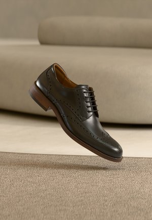 Sort læder broguesko med træsål, detaljerede perforeringer og snørebånd, der svæver over beige tæppe i et minimalistisk rum.