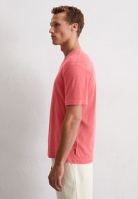 T-shirt en coton corail, col rond, manches courtes, avec un détail de fente sur le côté et une coupe droite, associé à un pantalon en lin de couleur claire.