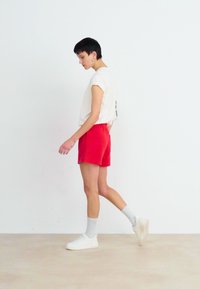 Maglietta bianca, pantaloni corti rossi e sneakers bianche. L'outfit presenta un design casual con una texture liscia e una palette di colori neutri.