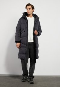 Manteau long et matelassé de couleur gris foncé avec une capuche, fermeture éclair et pressions, poches latérales et une finition texturée. Porté par-dessus une chemise blanche et un pantalon foncé.