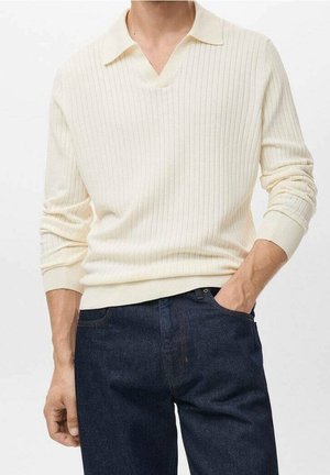 Mand iført cremefarvet ribbet langærmet sweater med krave og mørkeblå jeans, den ene hånd i lommen og den anden hængende ned.