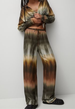 Osoba w luźnych spodniach tie-dye i pasującej zapinanej na guziki koszuli w zielonych, brązowych i żółtych tonach, stojąca na tle gładkiej białej ściany.