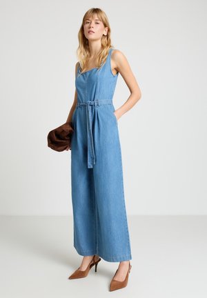 Donna che indossa una tuta in denim senza maniche con cintura e tacchi alti marroni, tiene in una mano una giacca marrone.