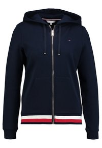 Sudadera con cremallera de color azul marino, con bolsillos frontales, capucha con cordón, y dobladillo a rayas en rojo, blanco y azul marino, que presenta un pequeño logo en el pecho.