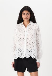 Hvit langermet skjorte med intrikate broderier og bølget kant, med knappestolpe foran og krage. Kombinert med svarte shorts.