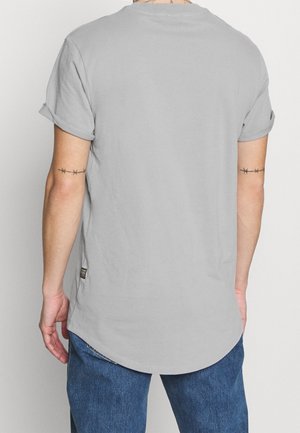 Šedé tričko s krátkými rukávy, s ohrnutými manžetami, zakřiveným lemem a malou štítkem na boku. Tkanina vypadá měkká a hladká.