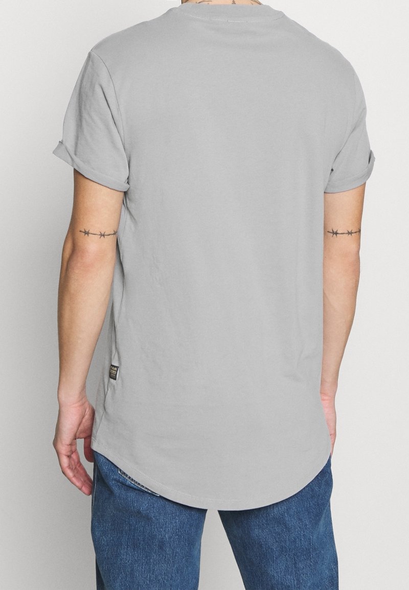 T-shirt gris à manches courtes avec poignets retroussés, ourlet incurvé et petite étiquette sur le côté. Le tissu semble doux et lisse.