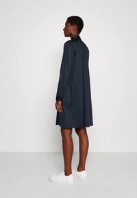 Navy blauwe jurk met kraag, lange mouwen en rugplooidetail, gecombineerd met witte sneakers. Glad materiaal, knielang ontwerp.