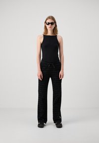 Svart ermeløs bodysuit, kombinert med løse sorte bukser med et gjentatt logo-mønster. Modellen har på seg svarte solbriller og mørke slip-on sko.