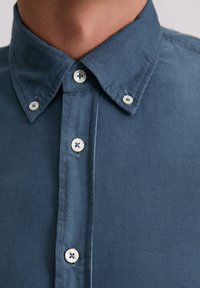 Blaues Cordhemd mit einem Knopfkragen, versehen mit hellen Knöpfen und vertikaler gerippter Struktur. Nahaufnahme des Stoffs.