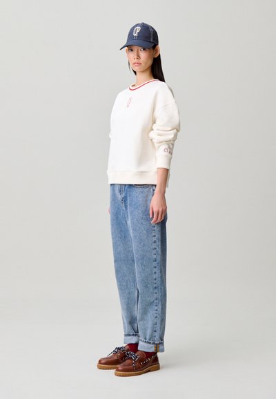 Sudadera blanca con detalles rojos, vaqueros azules y zapatos marrones con cordones azules. El modelo lleva una gorra azul marino con logo.