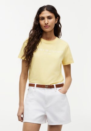 Kobieta o długich kręconych włosach, ubrana w żółtą koszulkę Tommy Hilfiger wpuszczoną w białe szorty z brązowym paskiem, stojąca z jedną ręką w kieszeni.