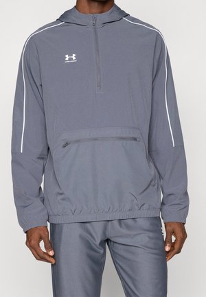 Hombre con un forro polar gris de manga larga Under Armour con cuello con cremallera, bolsillo delantero, ribetes blancos en las mangas y pantalones a juego.