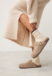 Sabots en daim beige avec une boucle latérale, assortis à des chaussettes crème côtelées et une robe tricotée beige clair. Les matériaux texturés sont visibles.