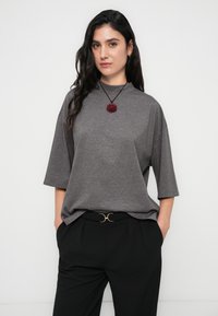 Γκρι φαρδύ μπλουζάκι με κοντά μανίκια και ψηλό λαιμό, συνδυασμένο με μαύρο παντελόνι που έχει ζώνη με χρυσές λεπτομέρειες και κόκκινο κρεμαστό κολιέ με λουλούδια.