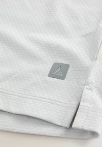 Jasnoszara koszulka sportowa z teksturowanej tkaniny, z małym szarym logo w kształcie trójkąta i wyszywanymi detalami na brzegach.