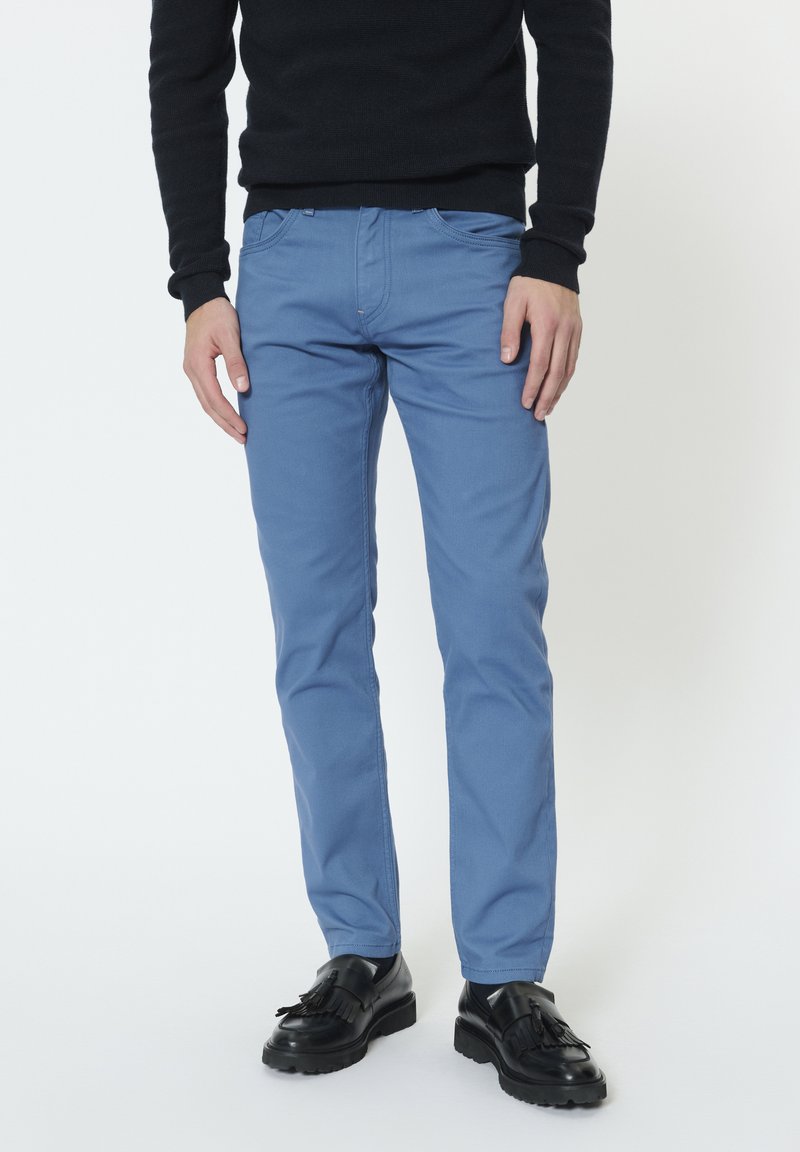 Férfi kék slim fit nadrágban, fekete pulóverben és fekete bojtos mokaszinban, egyszerű fehér háttér előtt áll.