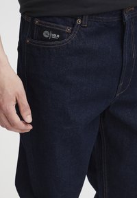 Temno modre kavbojke iz jeansa s krojem naravnost navzdol, okrašene s svetlo bakreno šivanko, etiketo na sprednjem žepu in teksturirano tkanino.