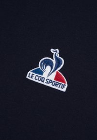 Le tissu bleu marine présente un logo brodé avec un coq stylisé et les couleurs du drapeau français. Le texte "LE COQ SPORTIF" figure sous le design.