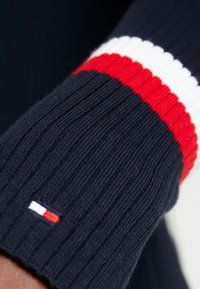 Manche de pull côtelé bleu marine avec des rayures rouge, blanc et rouge. Présente un petit écusson logo rouge, blanc et bleu au niveau du poignet.