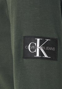 Mörkgrön texturerad tyg med en rektangulär svart etikett med vit text "CALVIN KLEIN JEANS" och en stiliserad "CK"-logotyp.