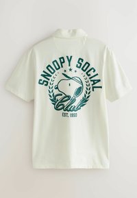 Korte mouwen polo shirt in crèmekleur, met een groene grafische afbeelding van Snoopy en de tekst "Snoopy Social Club" en "Est. 1950". Zacht materiaal.