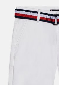 Pantaloni di cotone bianchi con tasche laterali diagonali, abbinati a una cintura intrecciata con strisce blu, rosse e bianche e una fibbia nera.
