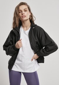 Svart lett jakke med hette, elastiske mansjetter og glidelås. Brukt over en hvit skjorte, sammen med glinsende lilla leggings. Glatt tekstur.
