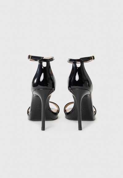 Sandales à talons hauts en cuir verni noir avec des brides de cheville et des talons aiguilles, présentées de dos sur un fond blanc uni.